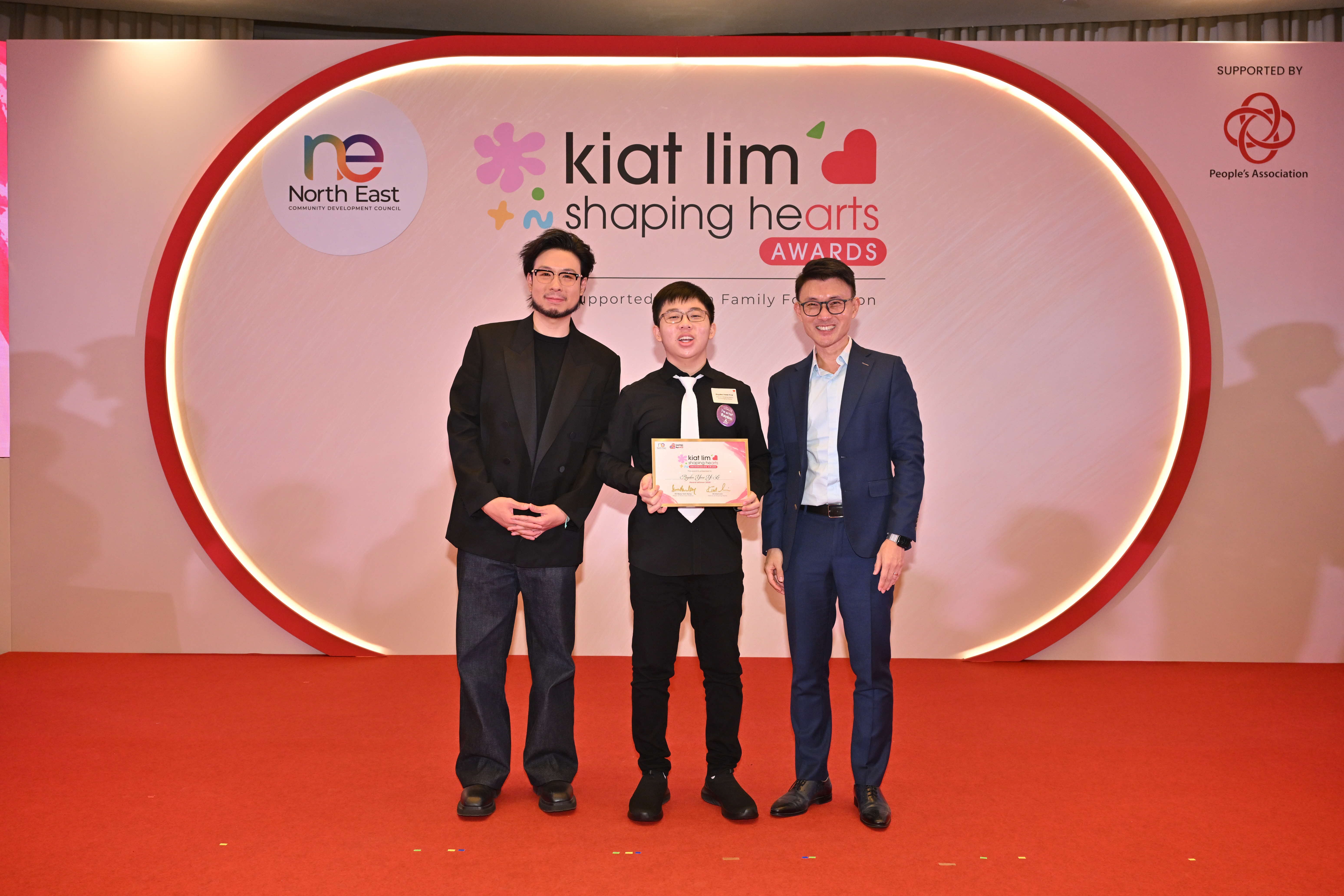 Award recipient standing on stage with the presenter, holding their certificate and posing for a group photo during the award ceremony, with a backdrop reading ‘Kiat Lim-Shaping Hearts Awards'.
